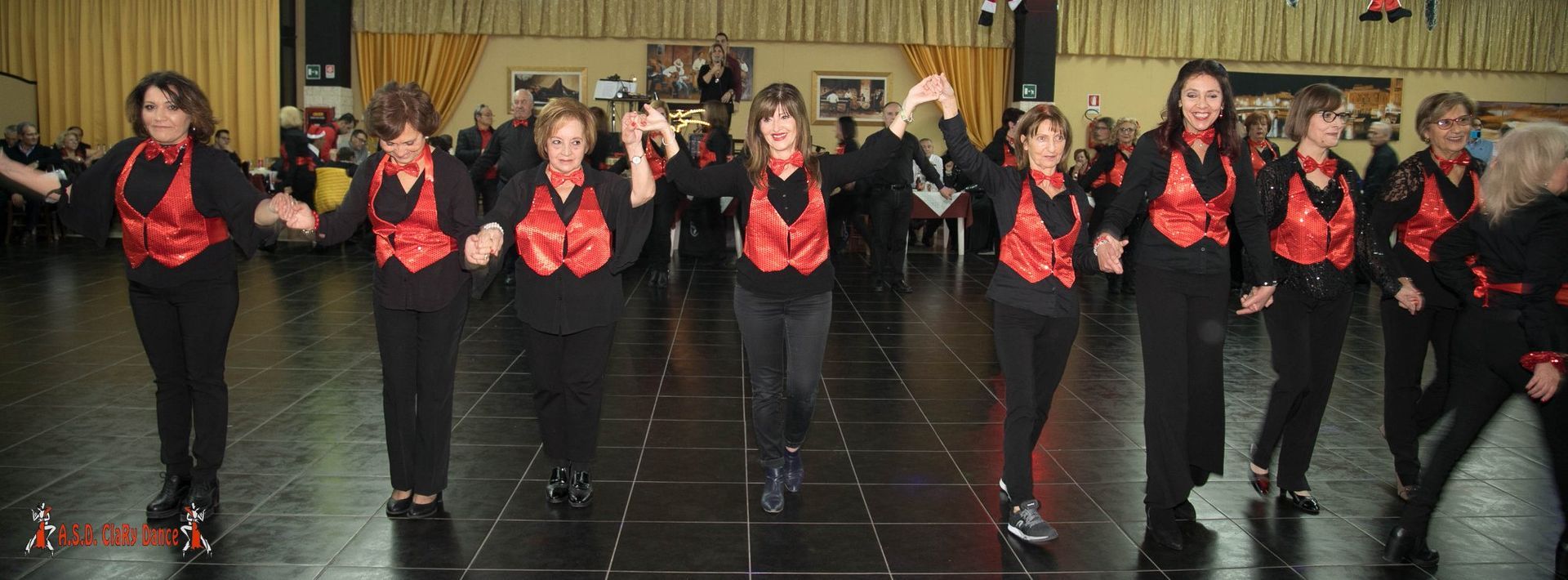 Un gruppo di donne con gilet rossi e abiti neri balla in una sala, con altre donne sullo sfondo.