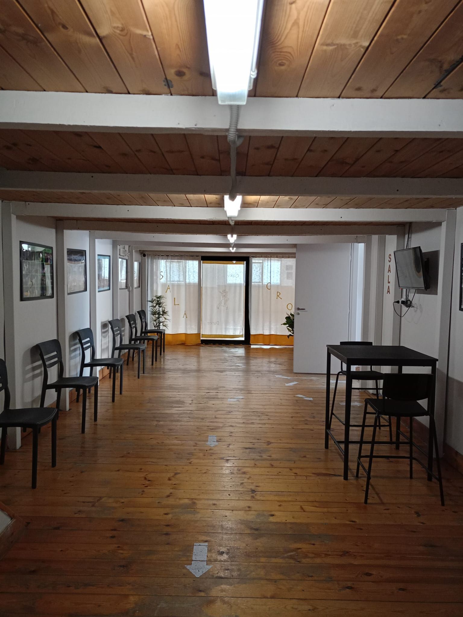 Una lunga stanza con pavimento in legno, file di sedie, tavoli e una porta in fondo. Luci fluorescenti e travi in ​​legno a vista.