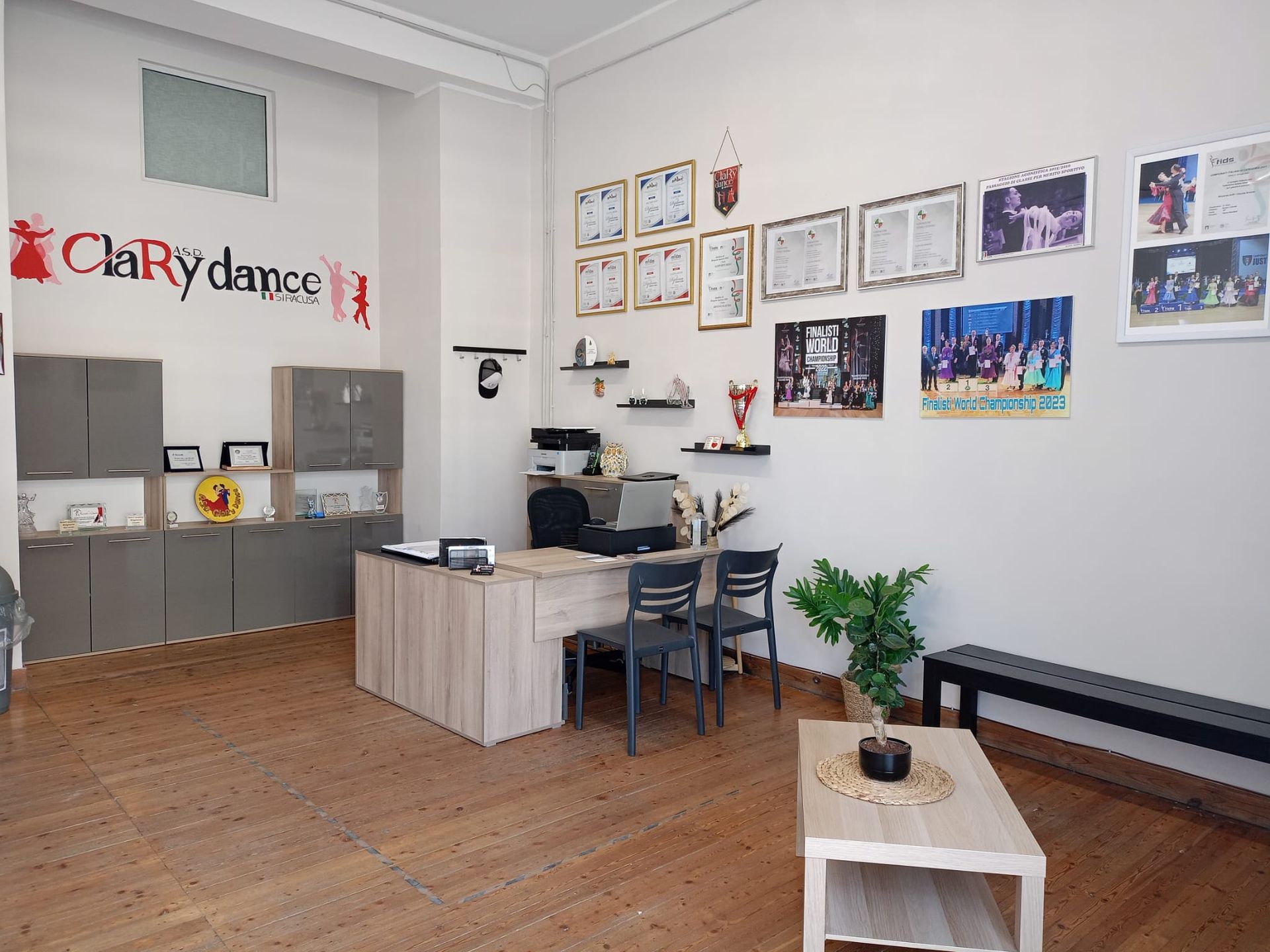 Interno dell'ufficio di uno studio di danza con scrivania, armadietti, premi e panca. Toni del grigio e del legno.