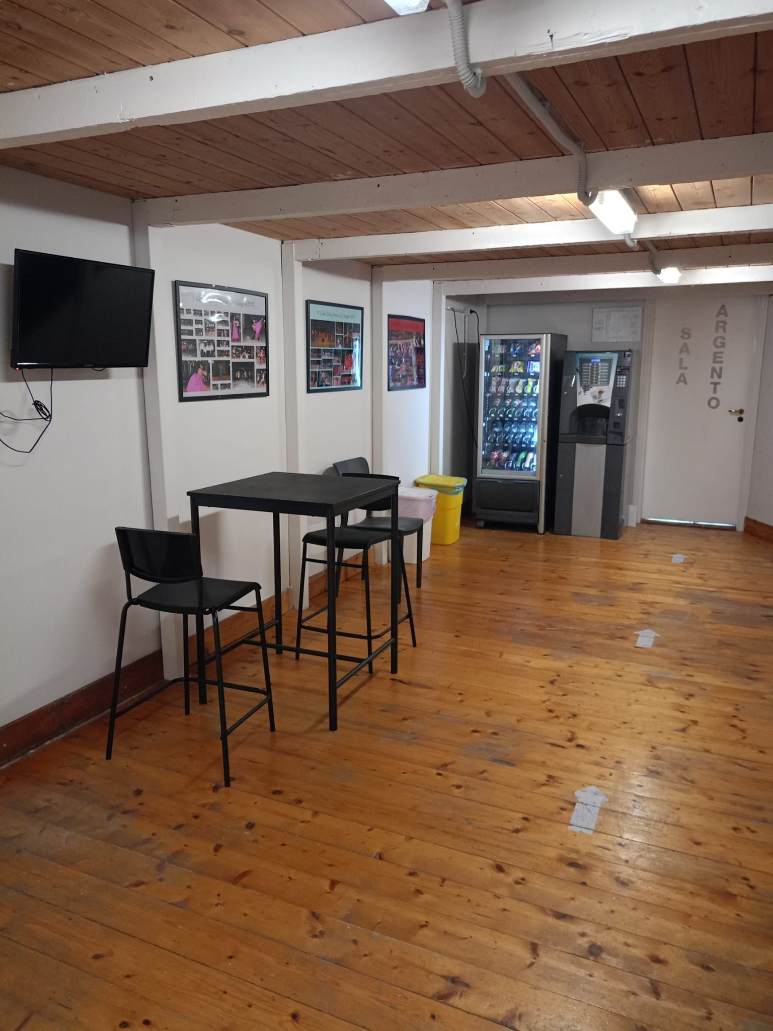 Una stanza con tavolo e sedie, distributori automatici e TV. Pavimento in legno e pareti bianche.