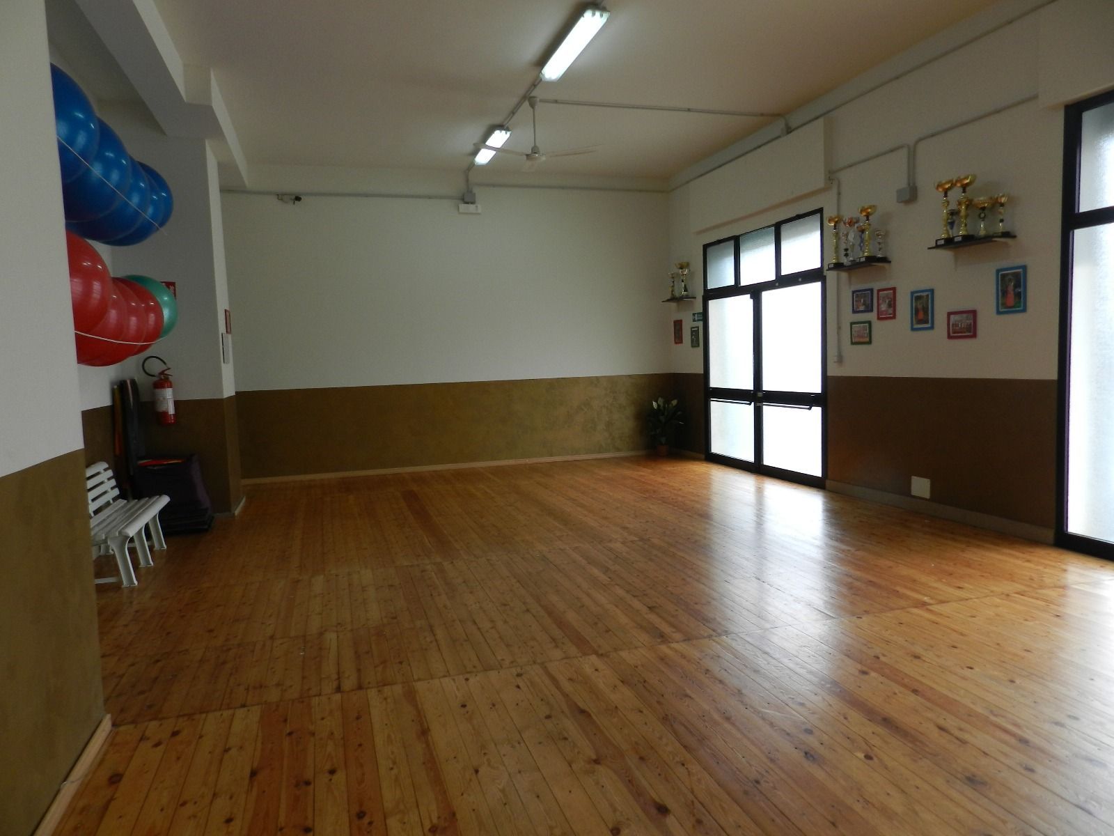 Stanza vuota con pavimento in legno, pareti bianche e ampie finestre. Sono visibili un estintore e dei palloncini.