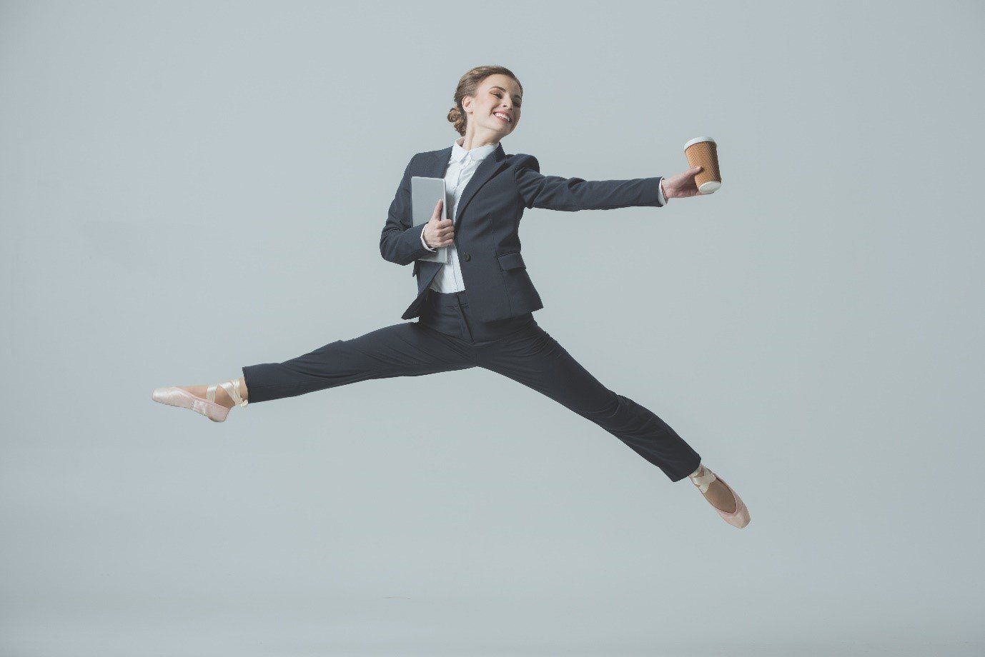 Female in dark business suit & ballet shoes leaping through the air with laptop and coffee in her hand