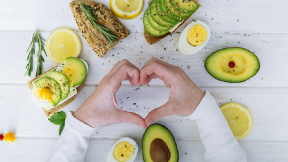 Hands making heart symbol with avocado, lemons, eggs and bread around it