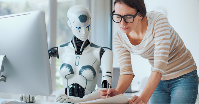Woman and a robot at a desk with a PC. The woman is writing on paper while the robot looks on
