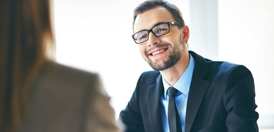 Man in suit sitting opposite a woman in a suit and having a chat. The man is smiling and engaged