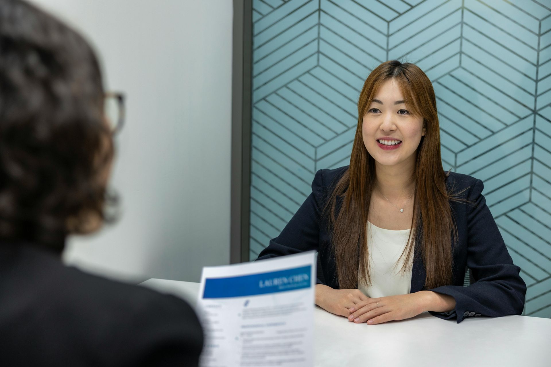 Woman in a navy blazer speaking across a desk in a modern office interview setting.