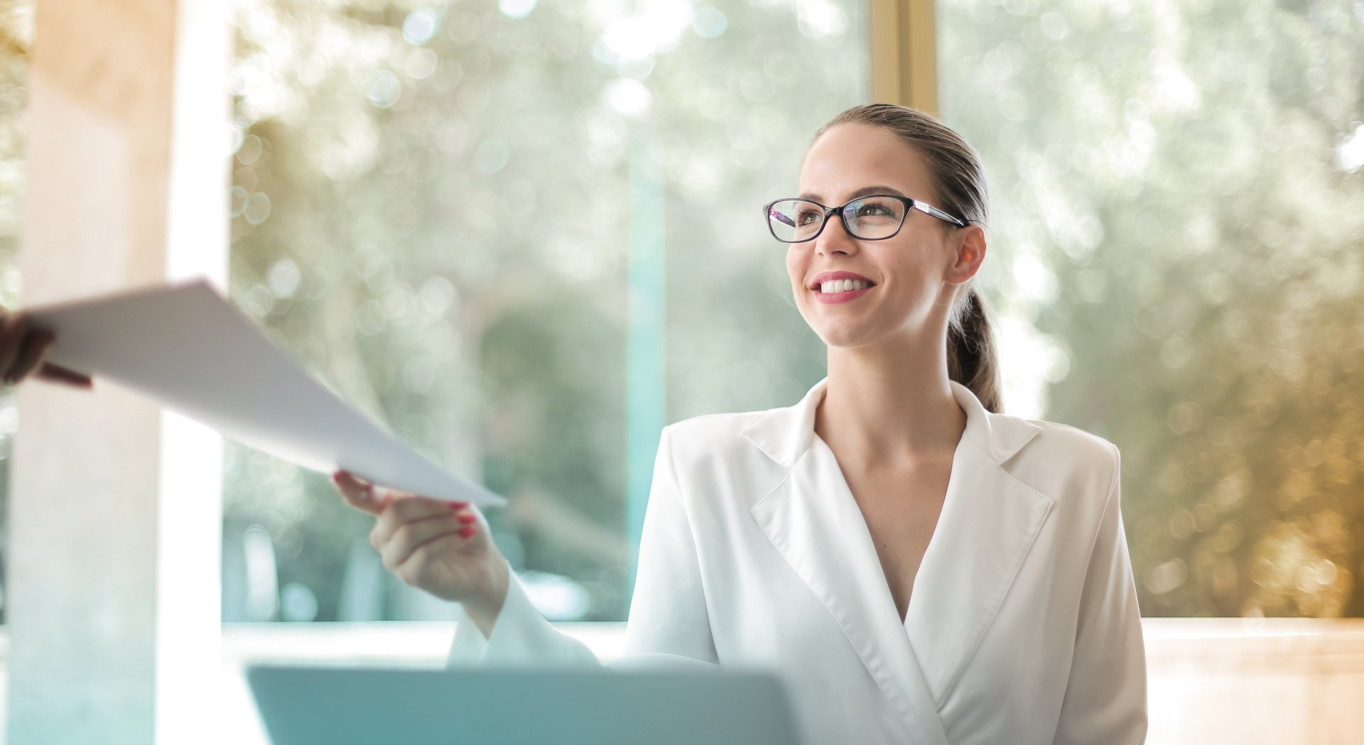 An smiling office worker sitting at their desk handing a bundle of papers to someone