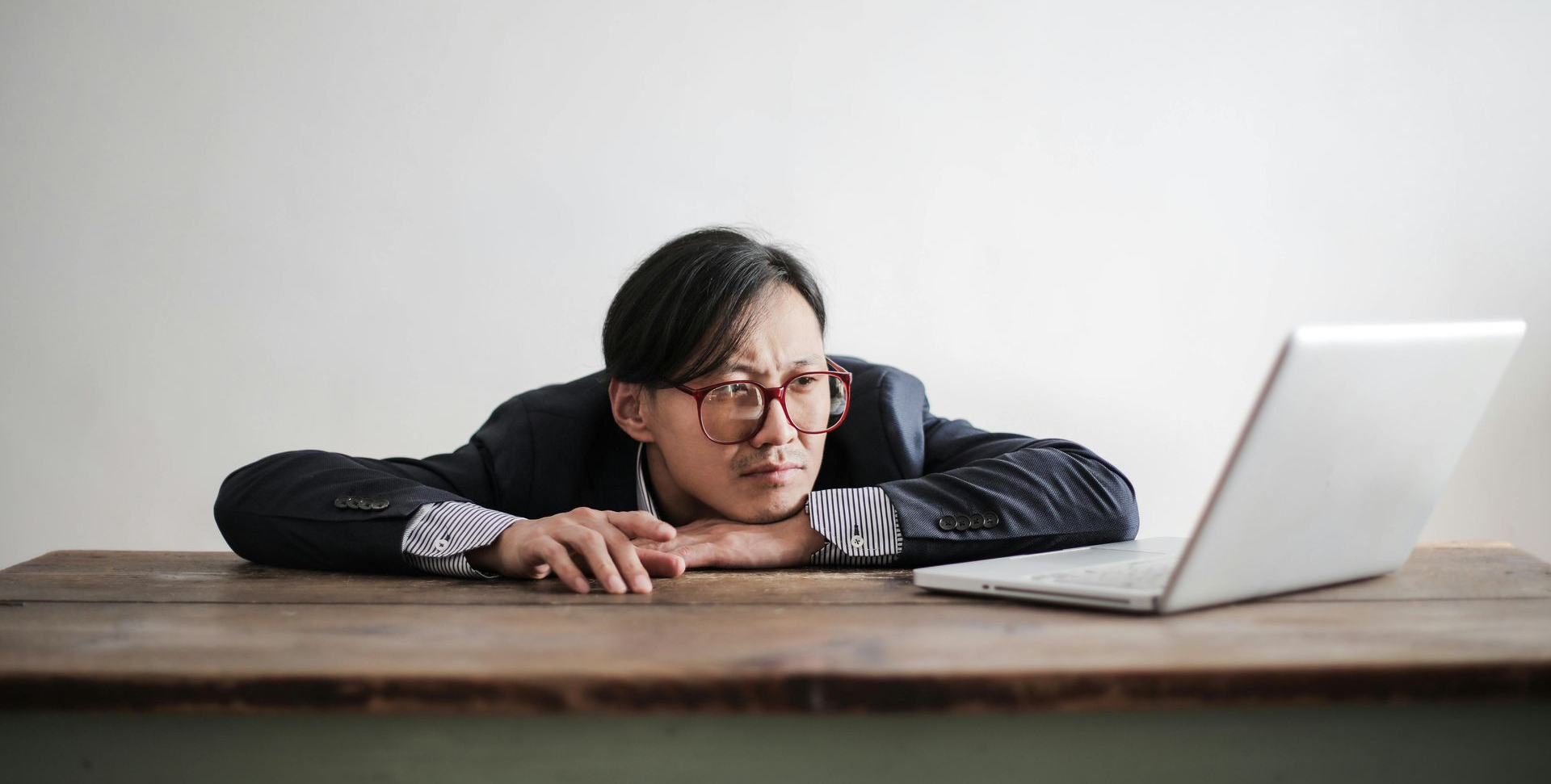 Man in a suit with his head resting on his hands and an unmotivated facial expression. He is sitting in front of his laptop. 