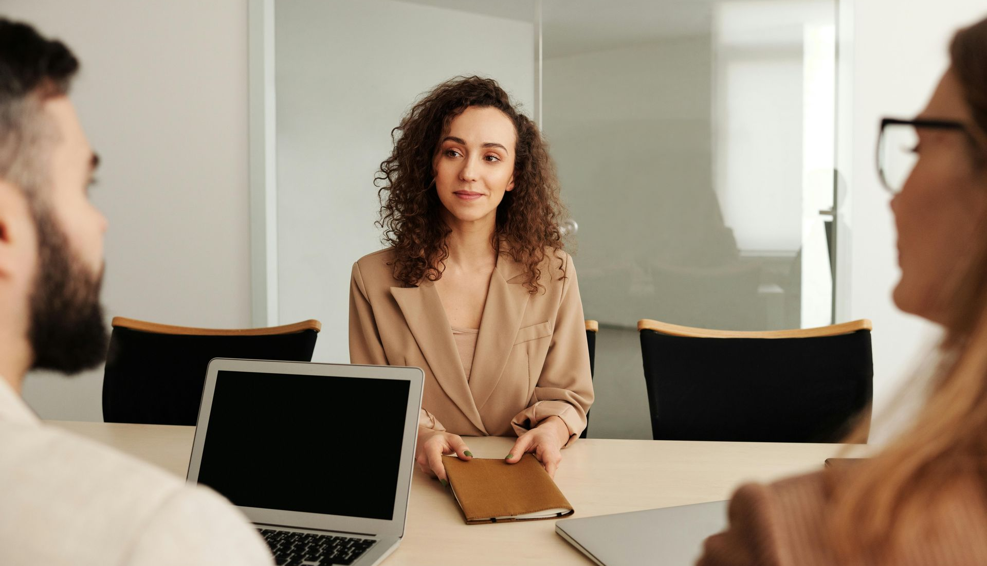Female sitting at an office desk with a notebook in her hand. There are two people sitting opposite her.