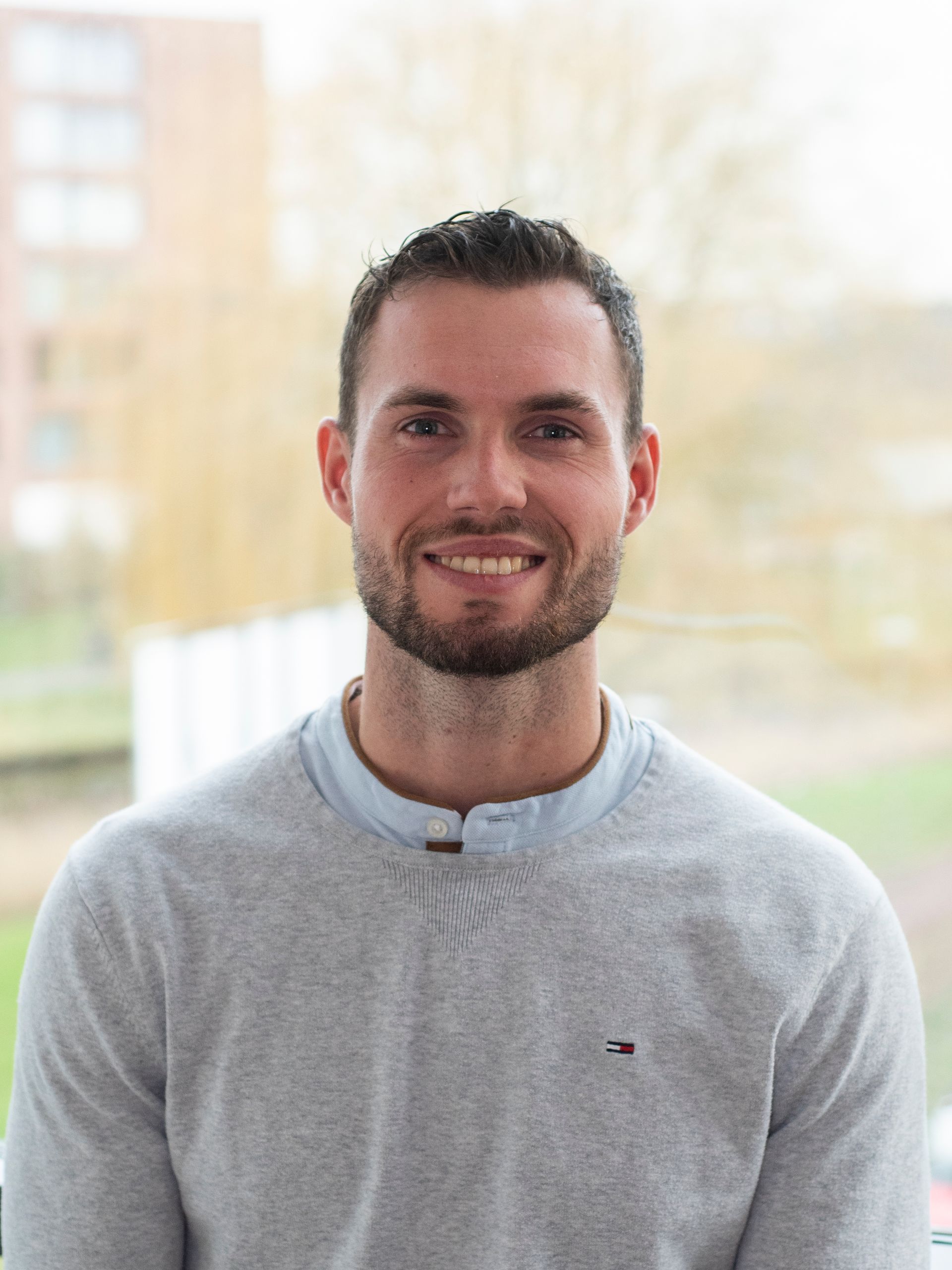 Een man met een baard draagt ​​een grijze trui en lacht naar de camera.