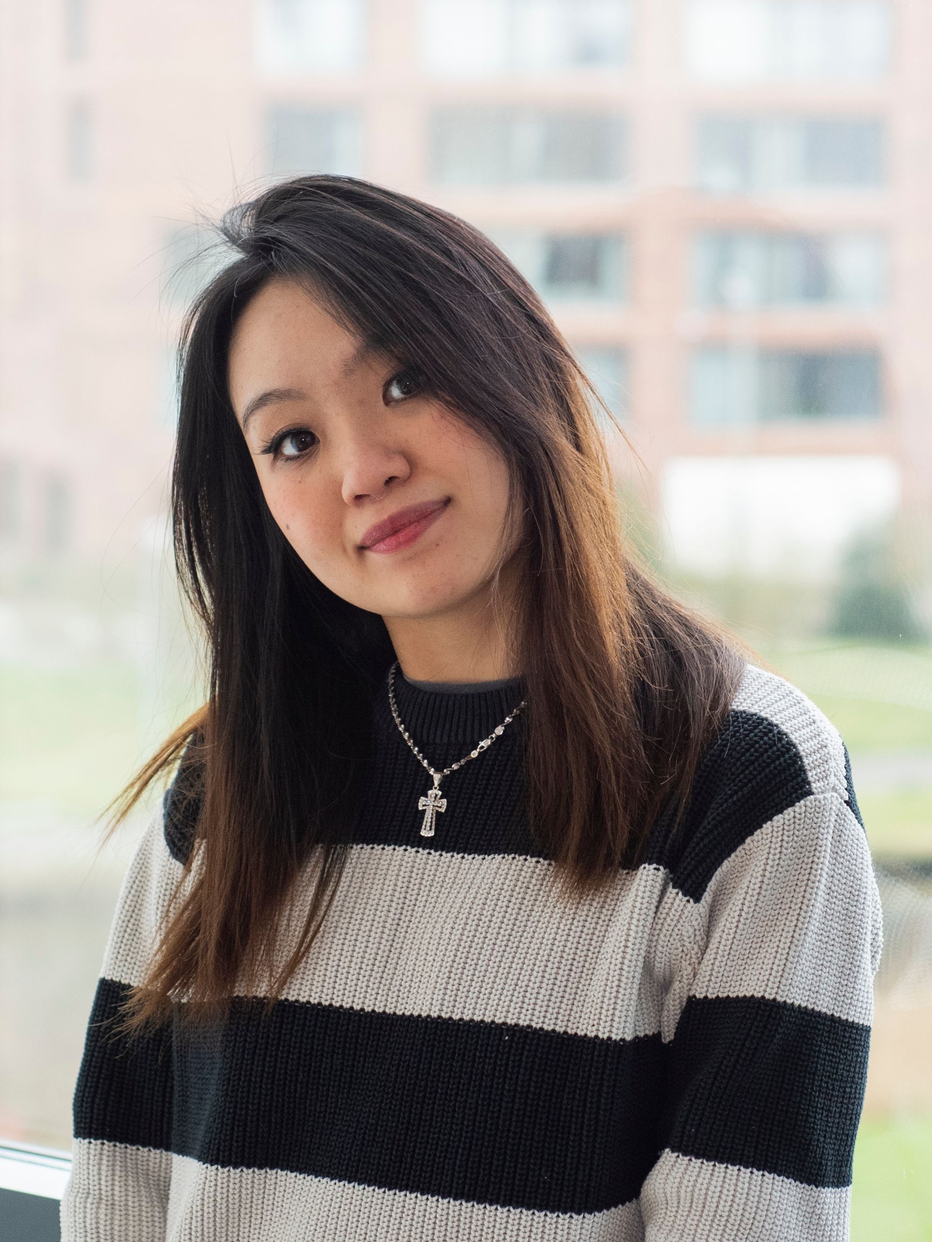 Een vrouw met een gestreepte trui en een ketting staat voor een raam.