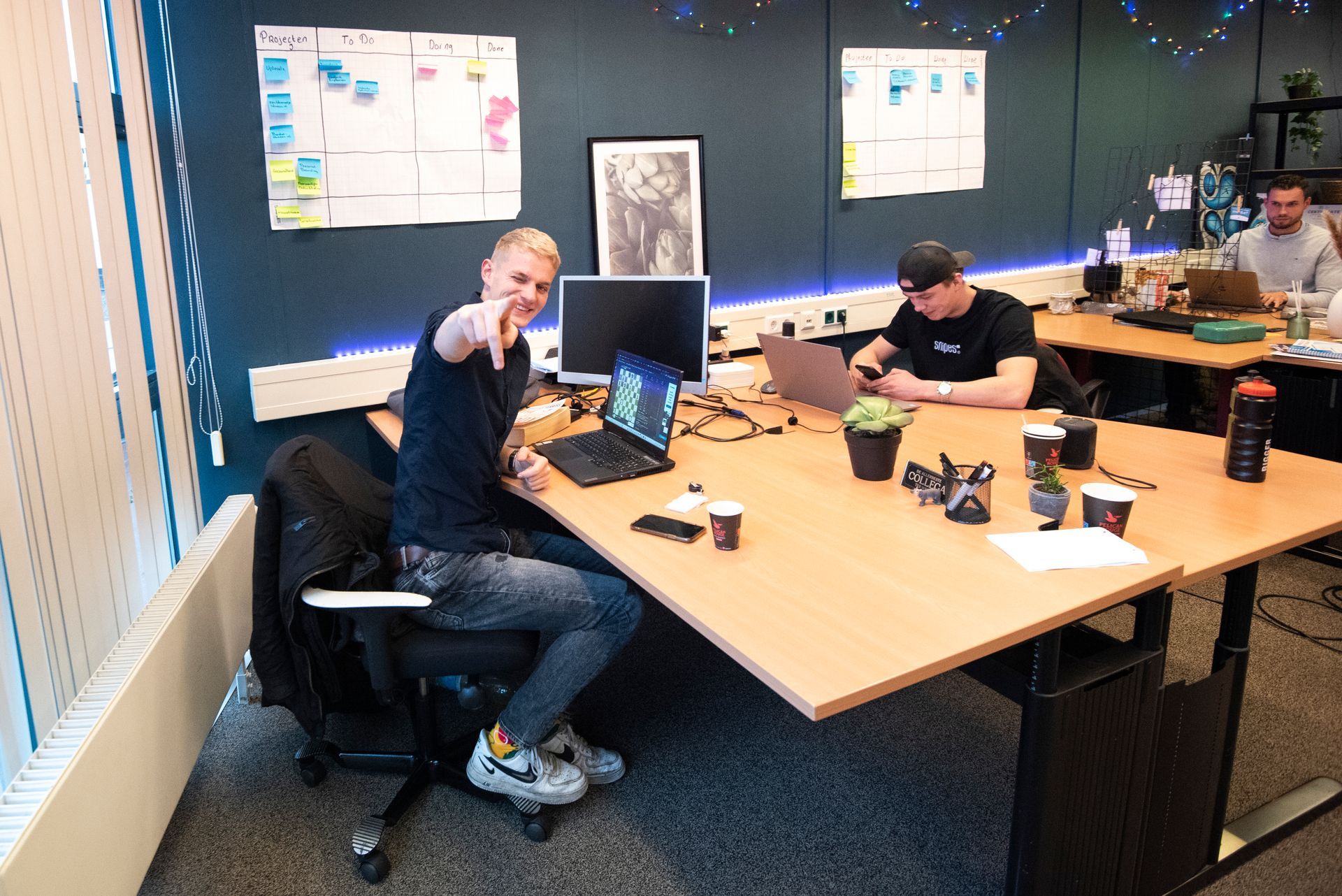 Een man wijst naar de camera terwijl hij op een kantoor aan een bureau zit.