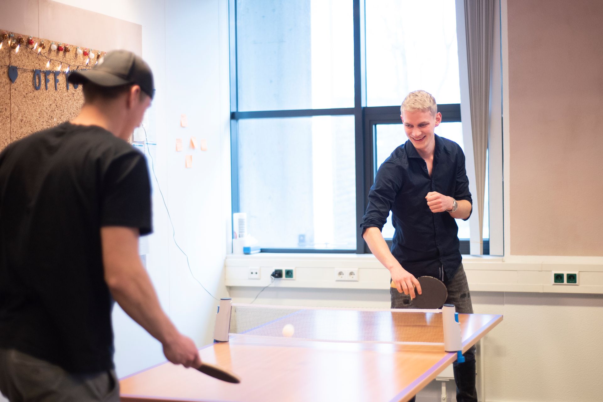 Twee mannen spelen pingpong op een tafel voor een raam.