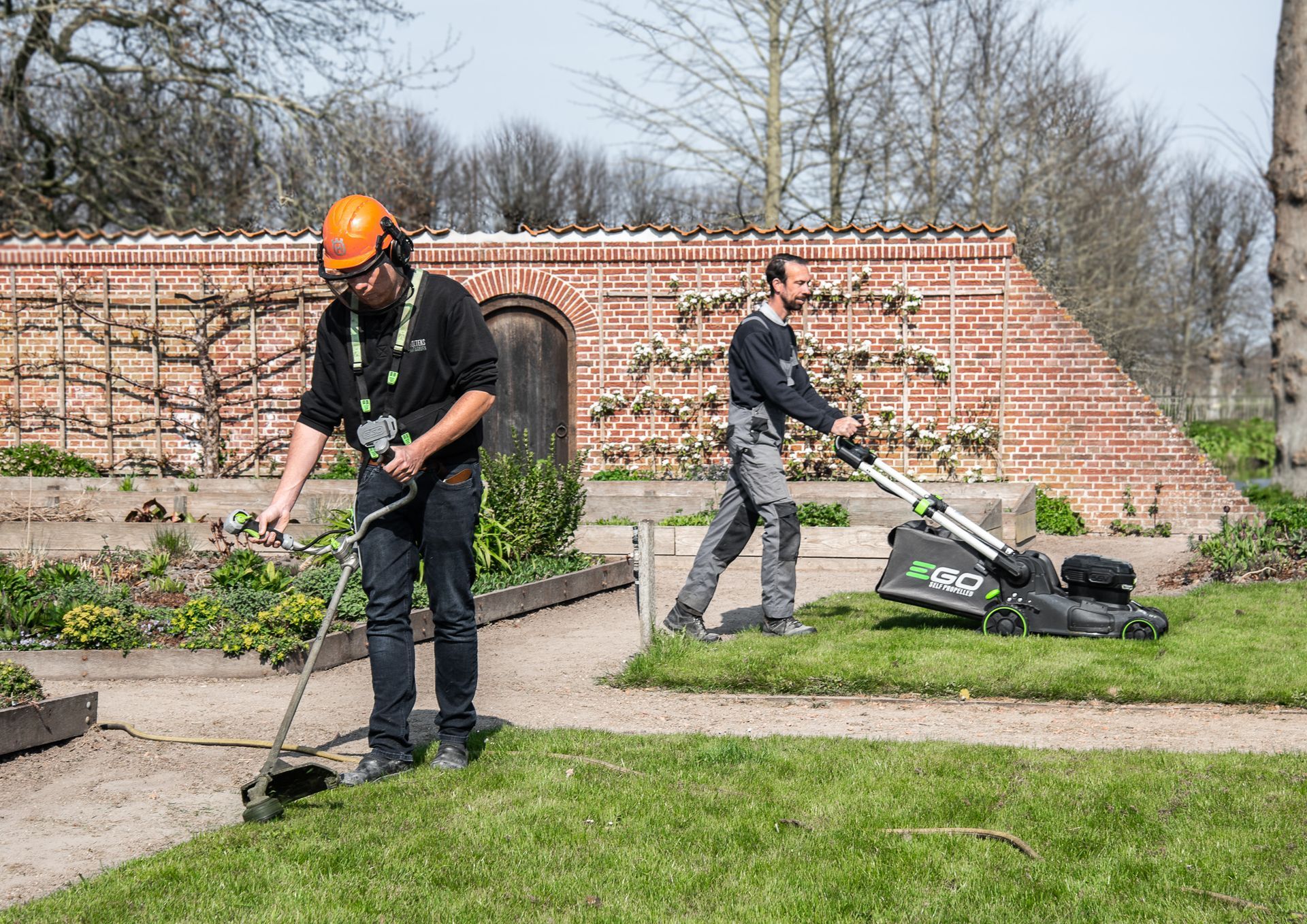 Twee mannen maaien het gras in een tuin met een grasmaaier.