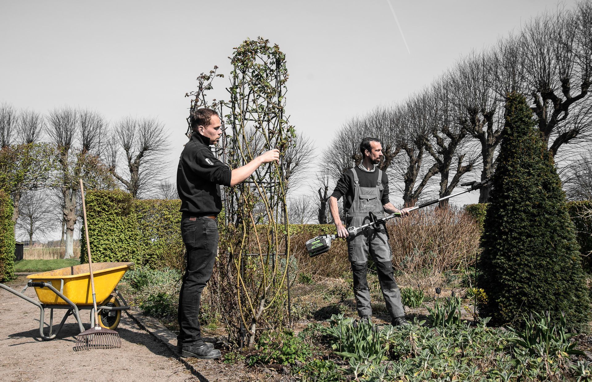 Twee mannen zijn in een tuin aan het werk met een kruiwagen.