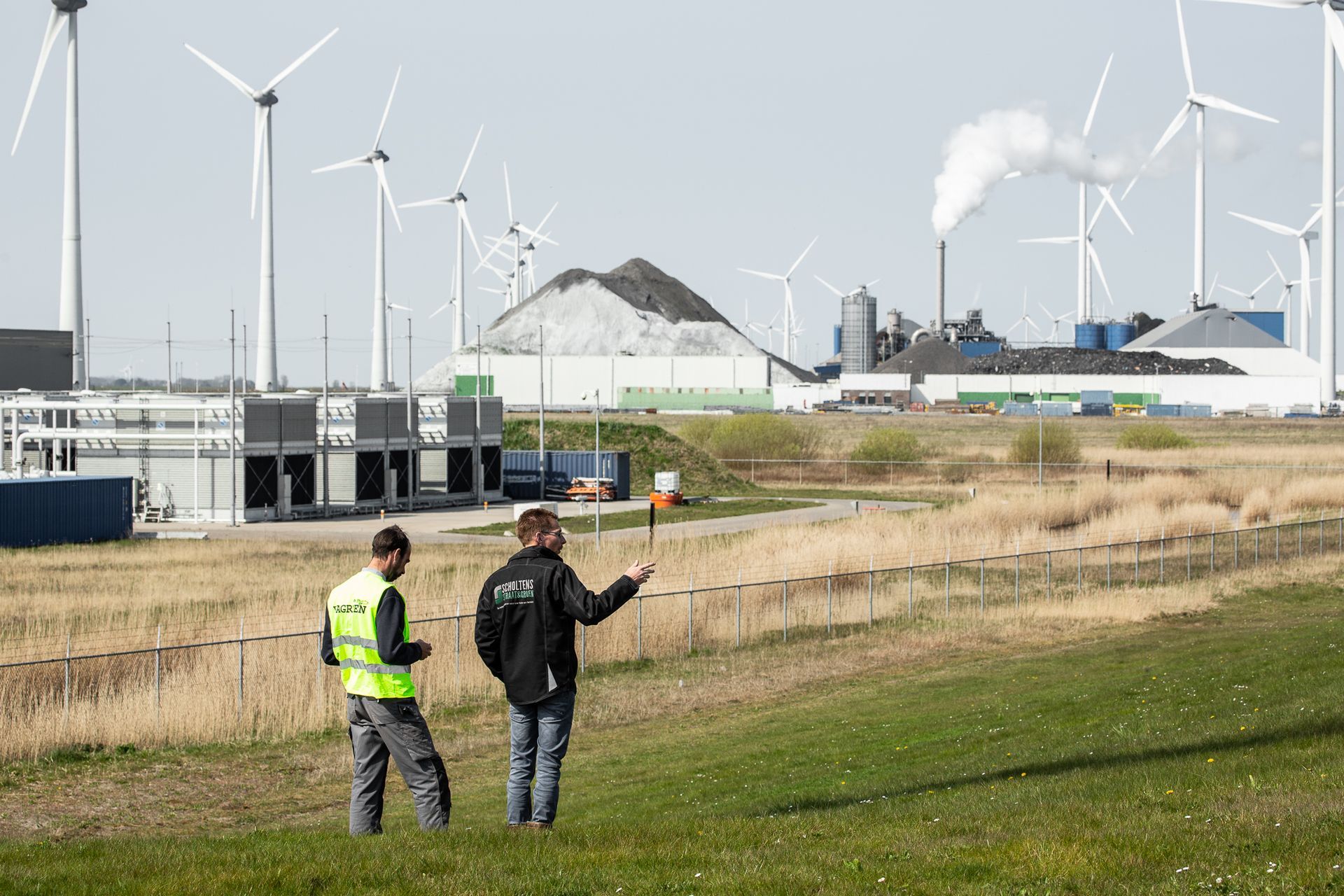 Twee mannen staan in een veld met windturbines op de achtergrond.