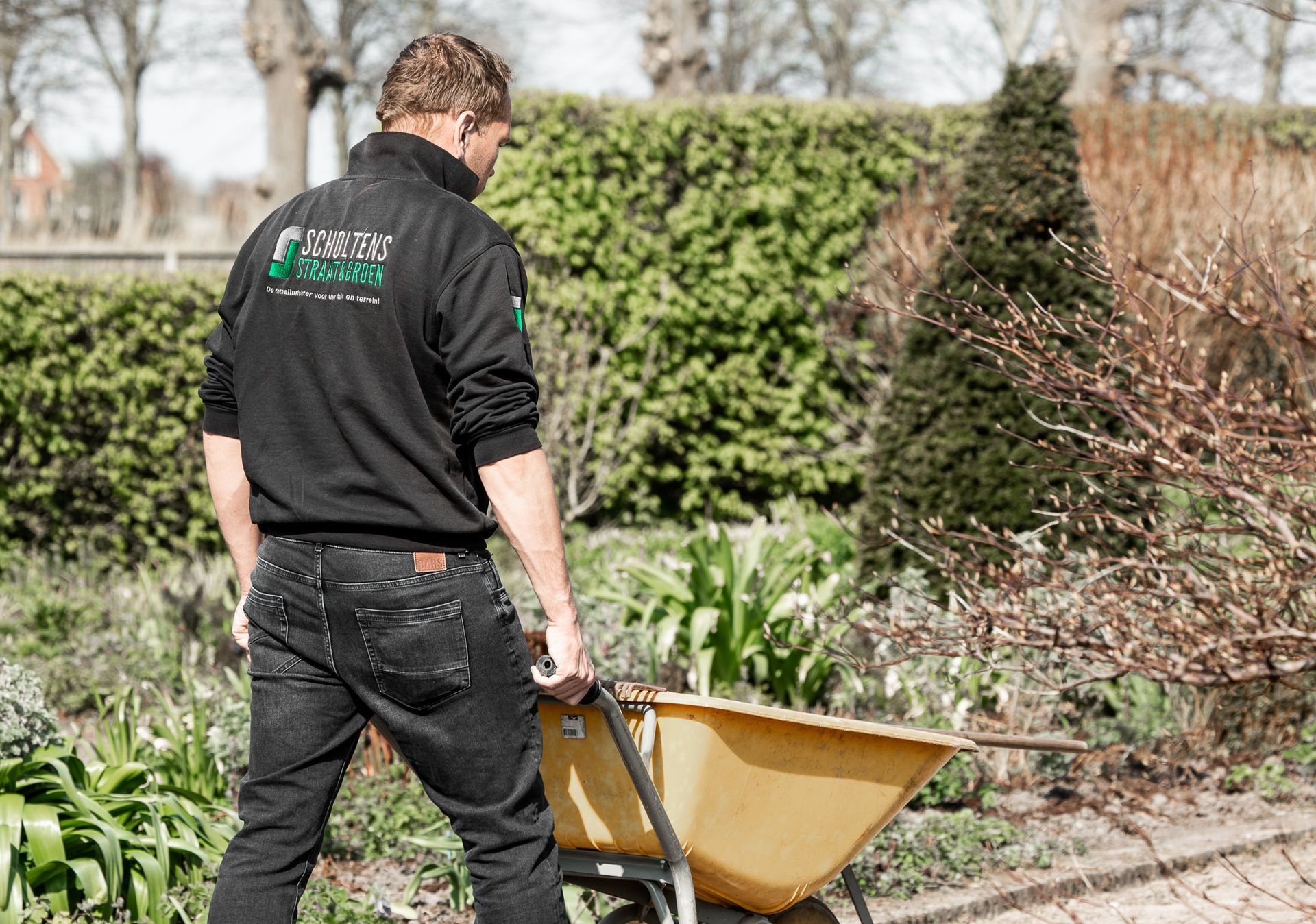 Een man duwt een kruiwagen in een tuin.