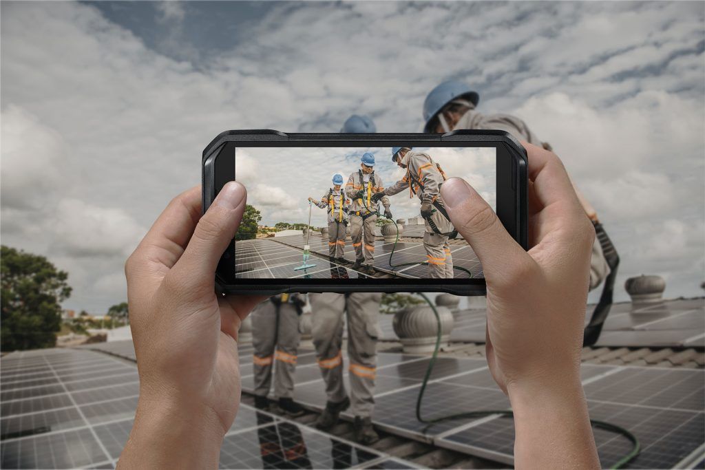 A person is holding a cell phone in their hands and taking a picture of construction workers on a roof.