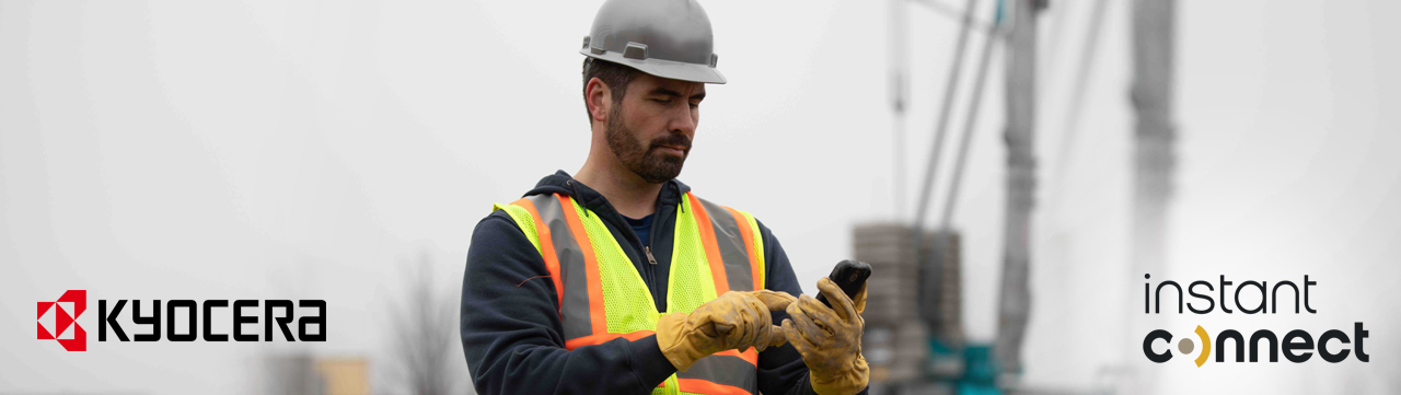 A construction worker wearing a hard hat and safety vest is looking at his cell phone.