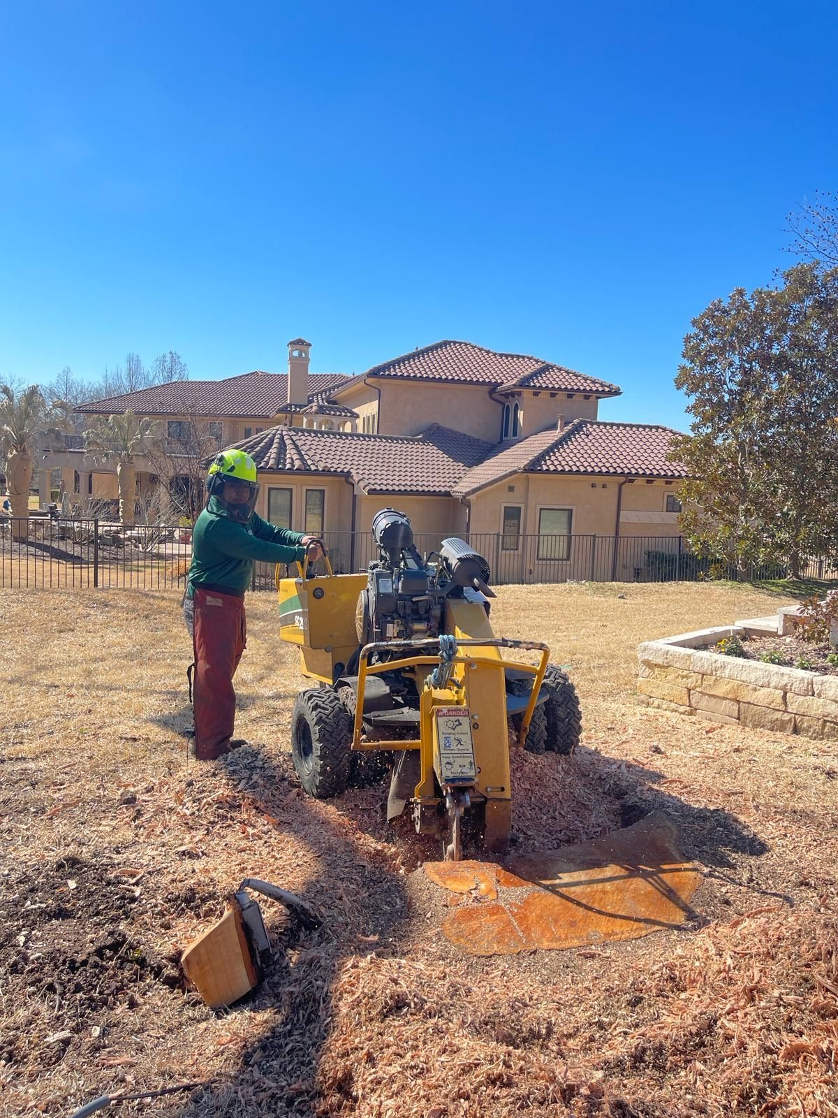 A man is working on a tree stump grinder in front of a large house.