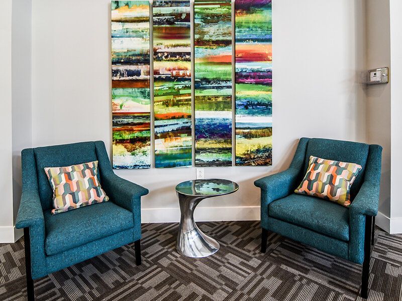 Two teal lounge chairs with patterned pillows and a glass side table in a modern community lounge.