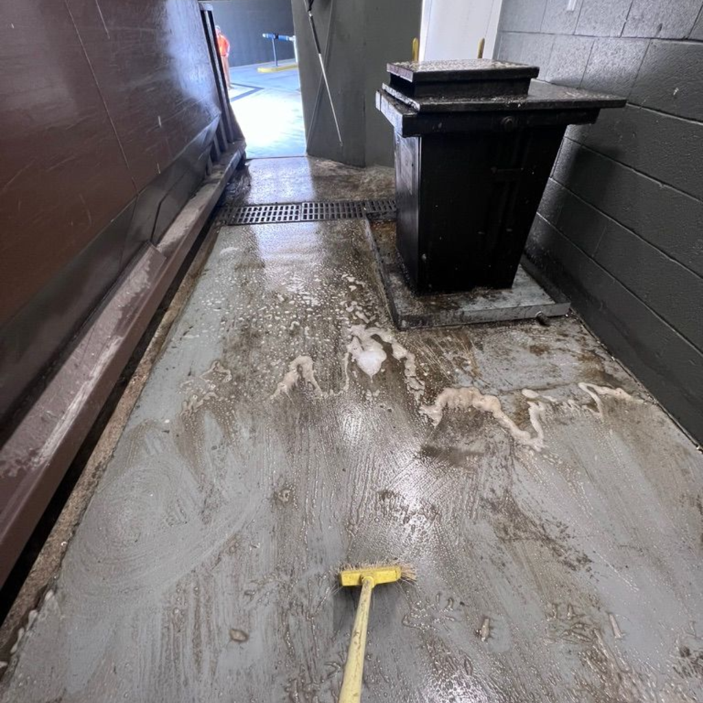 A broom and soapy water on a concrete floor next to a dumpster.