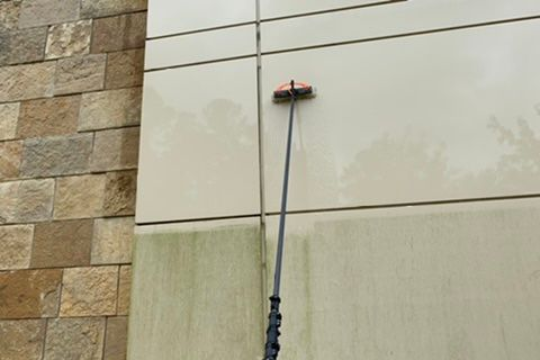A person is cleaning a building's exterior with a long-handled brush, removing algae from the light-colored wall.