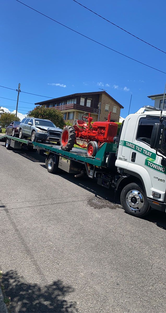 A Tow Truck is Carrying Two Cars and a Tractor Down a Street — Taree Tilt Tray Towing In Nabiac, NSW