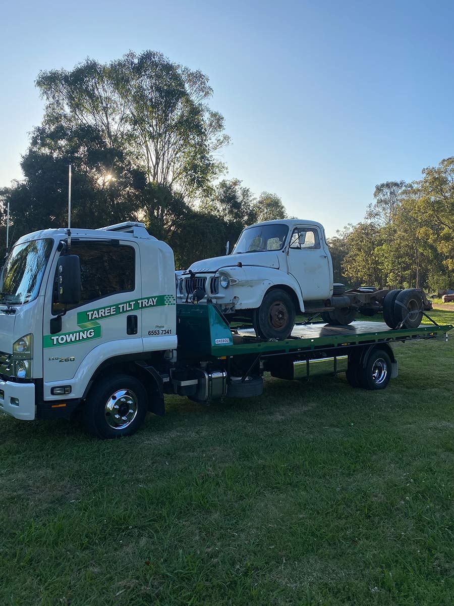 A Tow Truck is Towing a White Truck in a Grassy Field — Taree Tilt Tray Towing In Hallidays Point, NSW