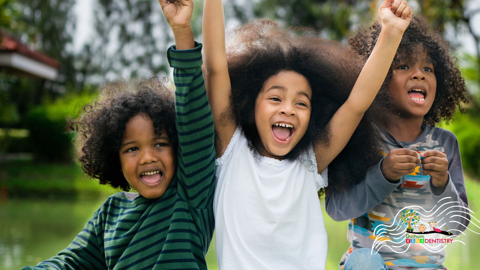 Three Black children cheering with arms raised, smiling by a pond.