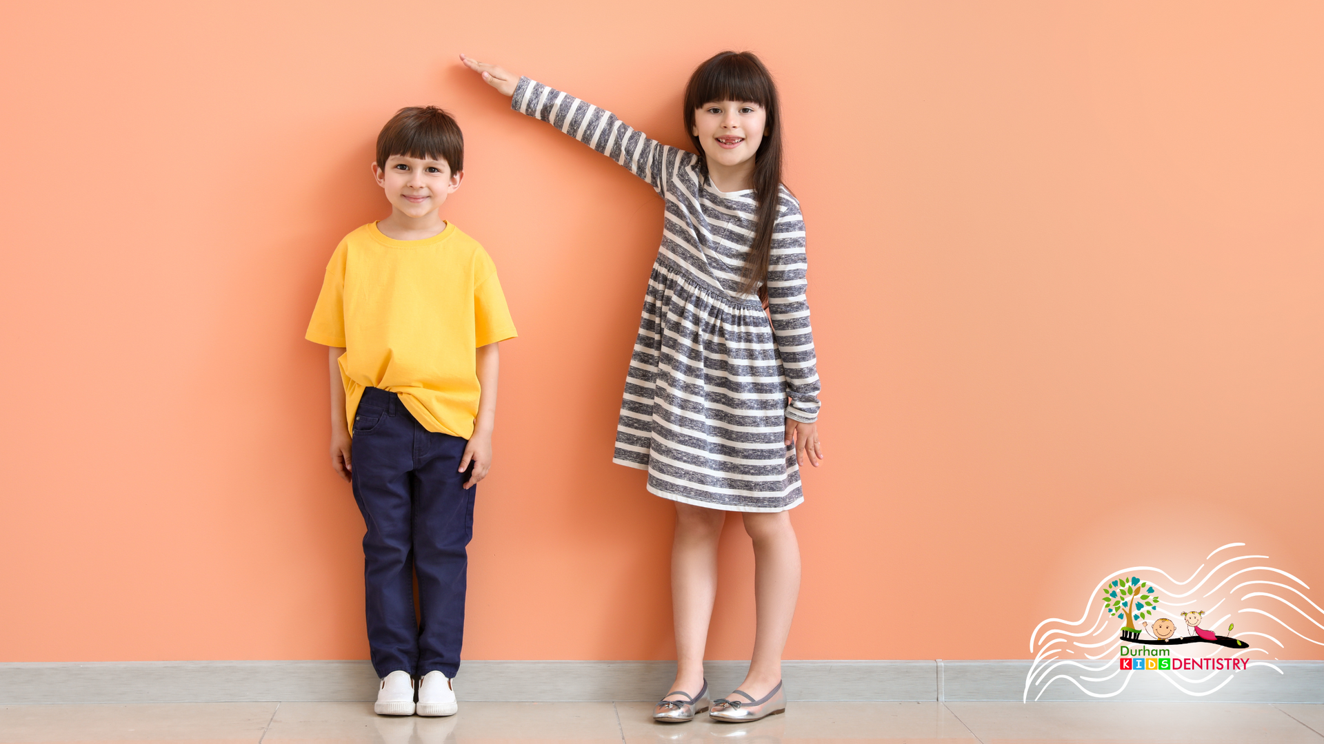 A boy and a girl are measuring their height against an orange wall.