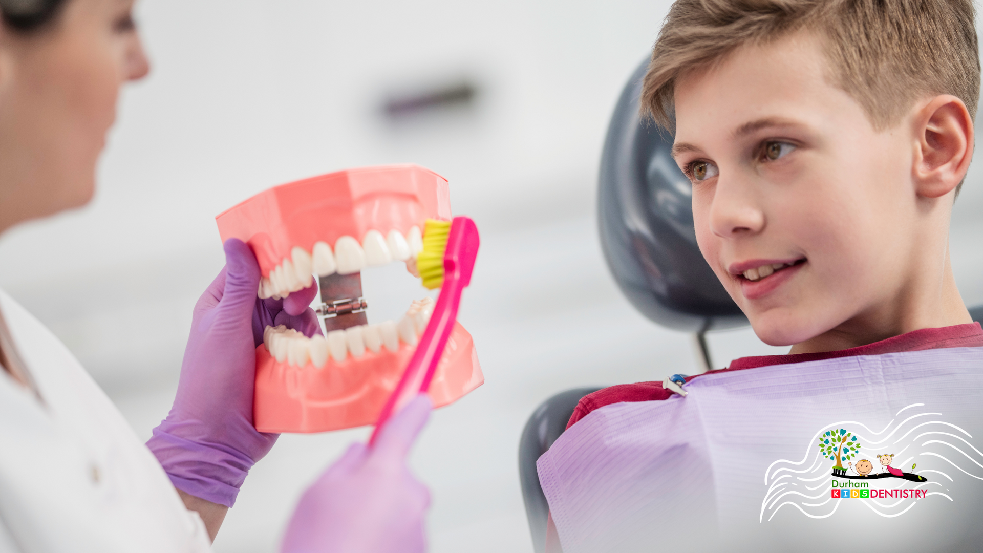 A young boy is sitting in a dental chair while a dentist shows him how to brush his teeth.