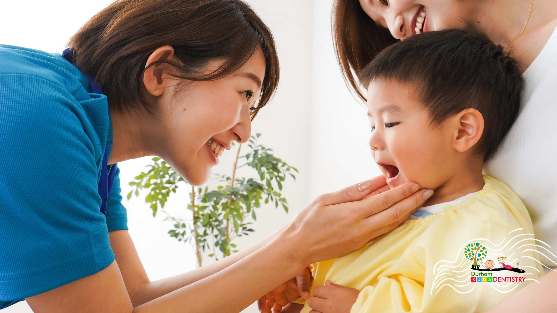 Smiling woman examining a toddler's mouth, another woman in the background, indoor setting.