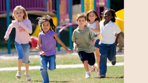 Five children running and smiling on a grassy playground with colorful equipment in the background.