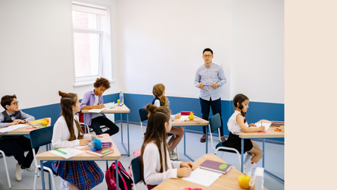 A teacher stands at the front of a brightly lit classroom as students sit at desks, working on their assignments.