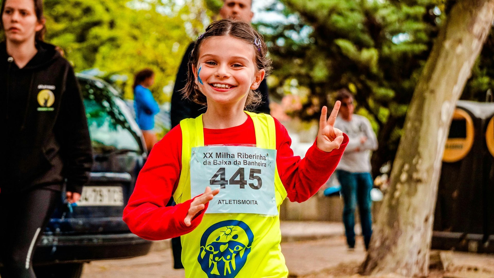 A young girl wearing a yellow vest with the number 445 on it is giving a peace sign.