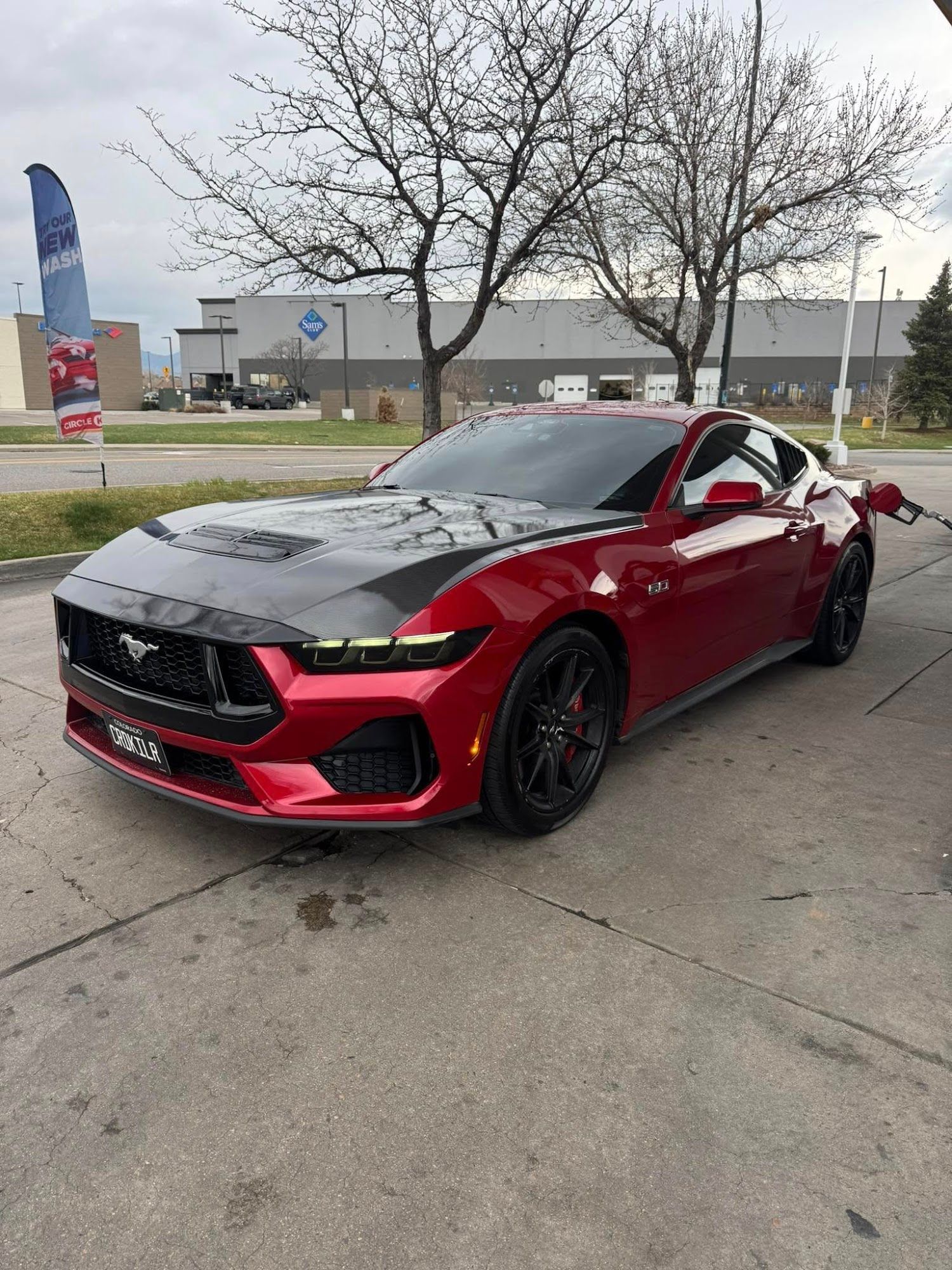 Red Ford Mustang coupe with carbon fiber hood, parked at a gas station.