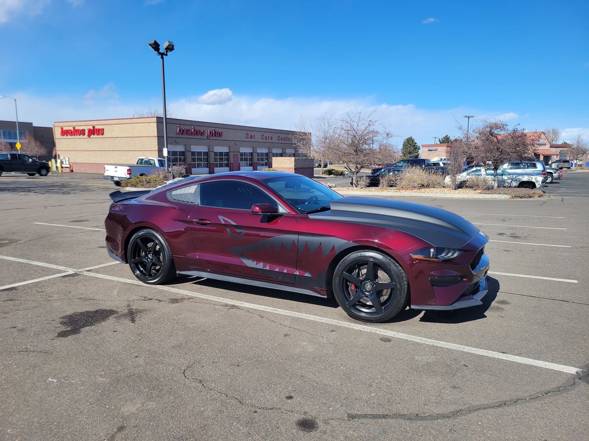 A maroon Ford Mustang parked in a lot, with a black hood and racing stripes; buildings in the background.