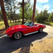 Red vintage Corvette parked on asphalt road in front of trees.