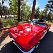 Red classic convertible car parked on a paved road, surrounded by trees and a sunny landscape.