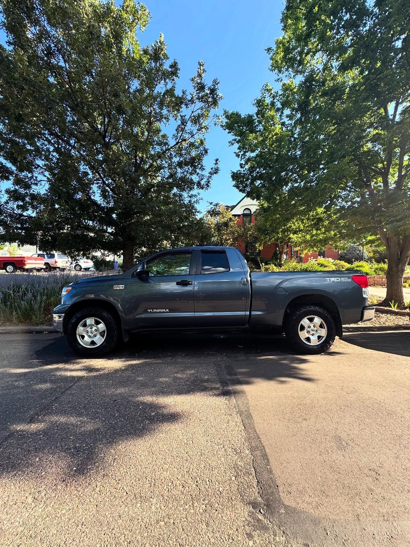 Gray Toyota Tundra pickup truck parked on asphalt street with trees and buildings in the background on a sunny day.