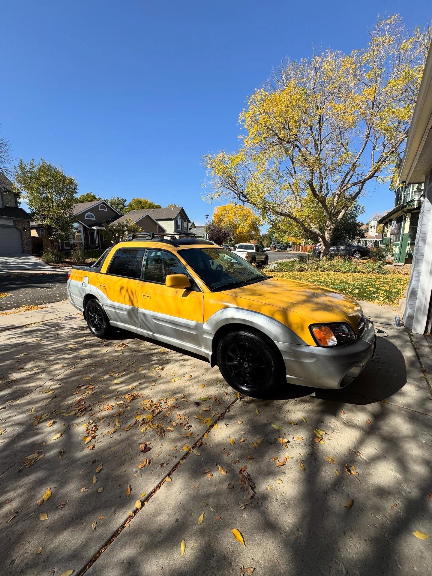 Yellow and silver Subaru Baja pickup truck parked on a driveway in front of a house, under a sunny sky.