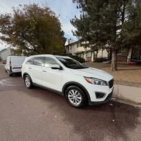 White SUV parked on a street with a van in the background. Buildings and trees are visible.