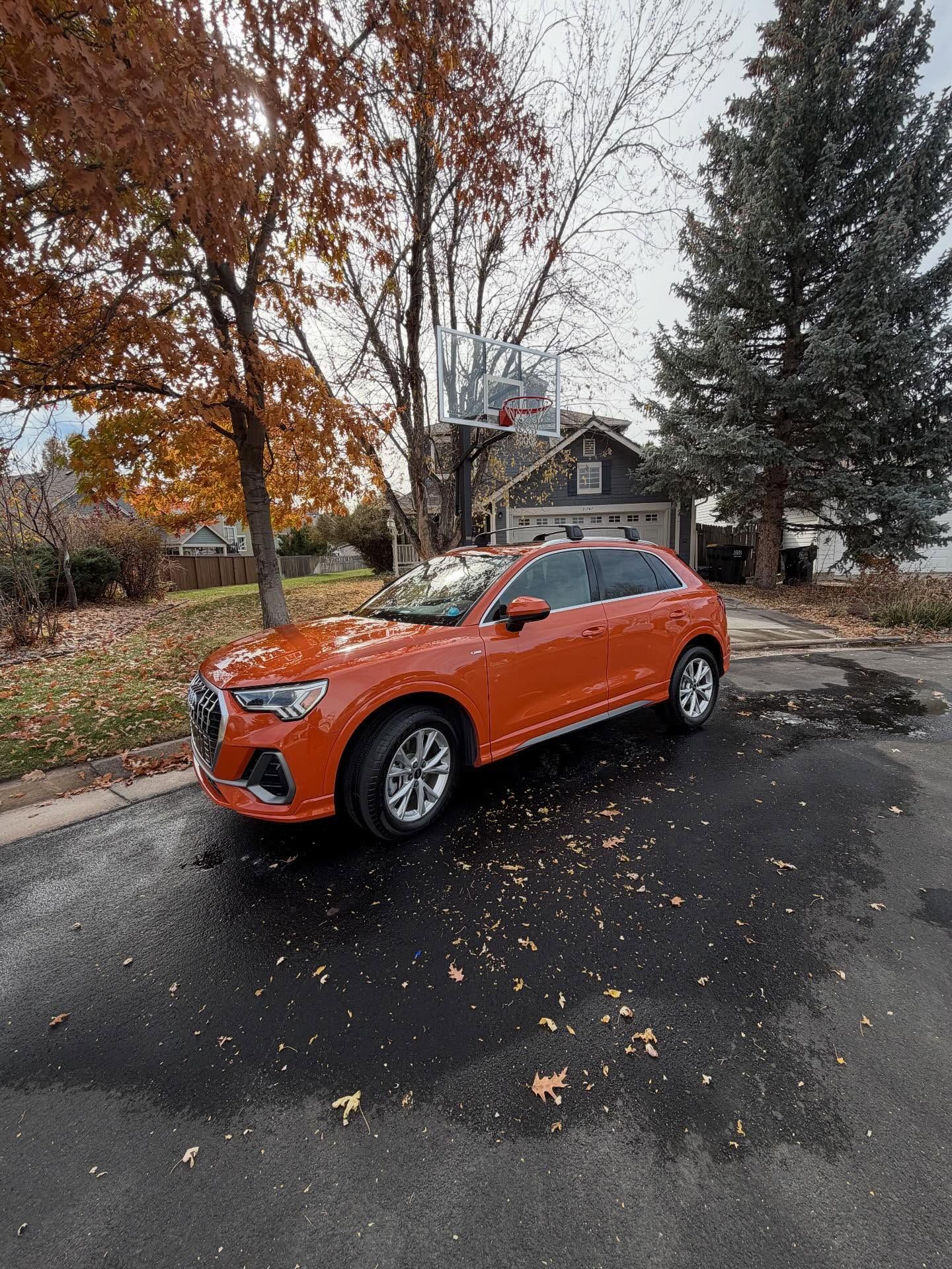 Orange Audi SUV parked on wet asphalt in front of a house with basketball hoop; fall foliage.