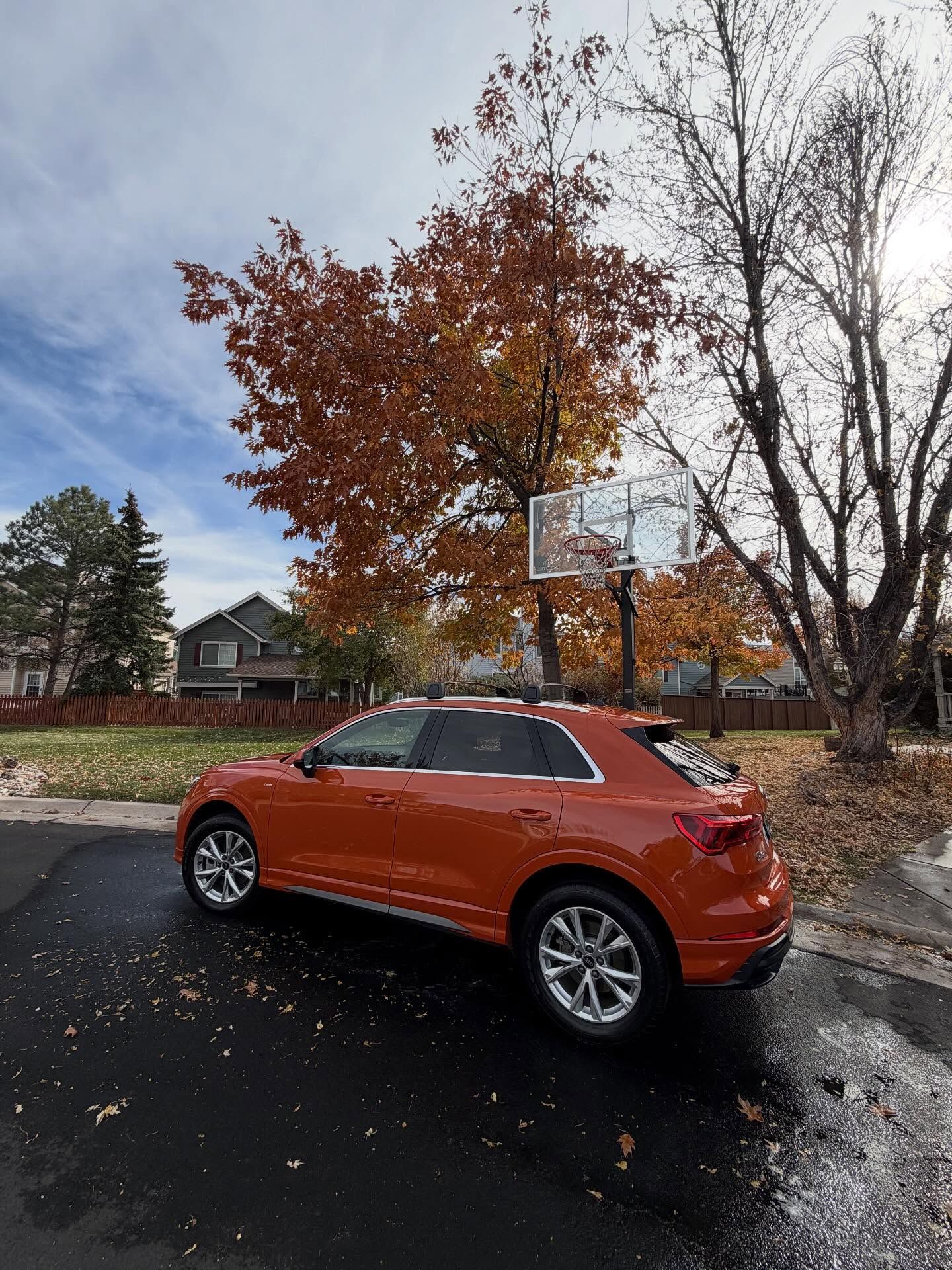 Orange SUV parked on wet asphalt in front of a tree with autumn leaves and a basketball hoop.