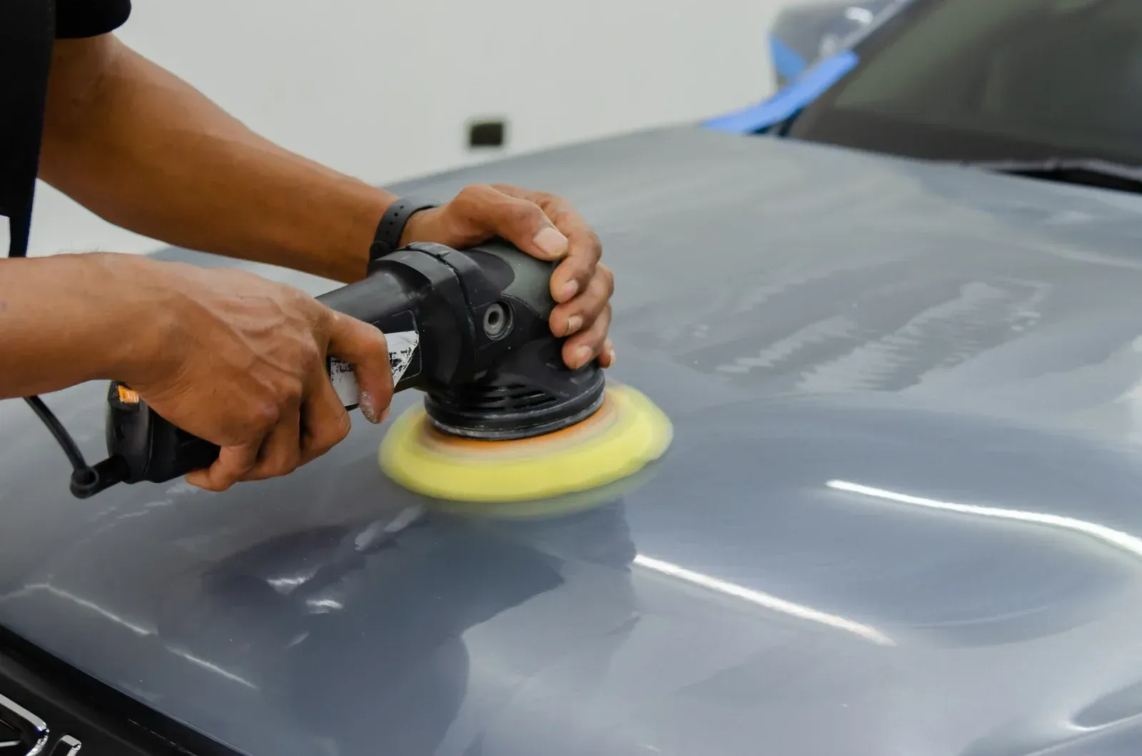 A person using a power polisher on a gray car hood. The polisher has a yellow pad.