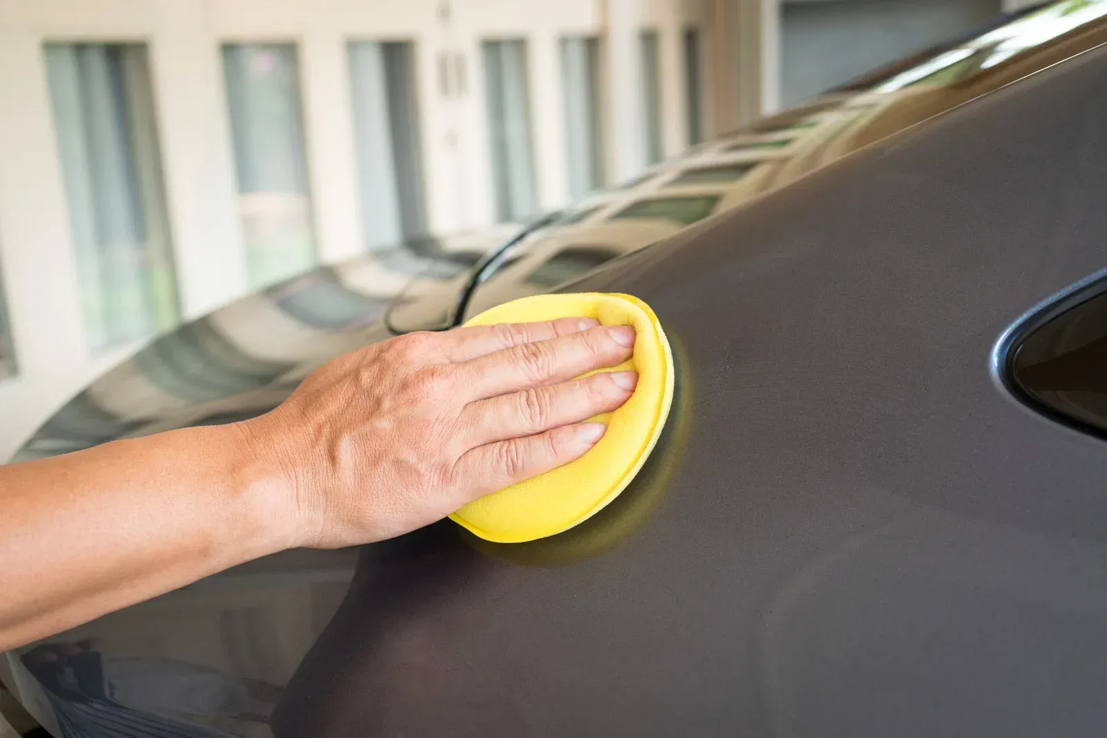 A person with arm tattoos washes a car covered in white foam with a red sponge.
