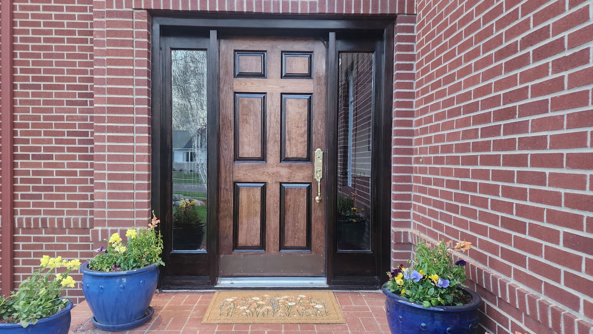 Wooden front door with sidelights, framed by red brick. Blue pots with flowers flank the entrance.