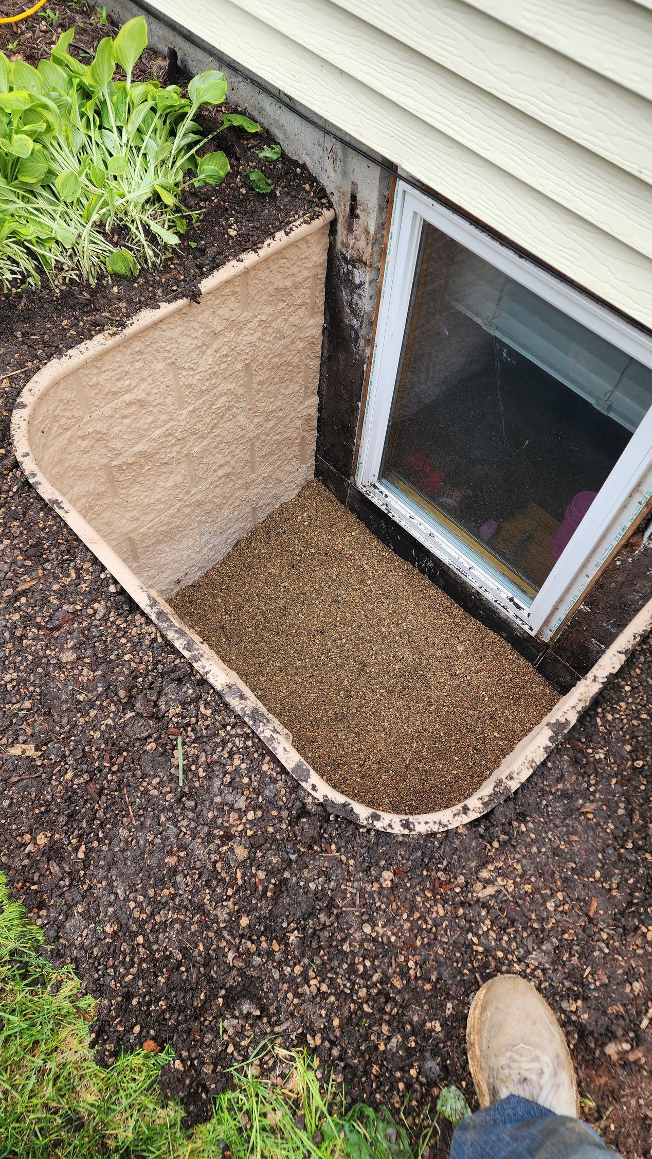 Window well with gravel, next to a home's foundation and window. Brown soil and green plants surround it.