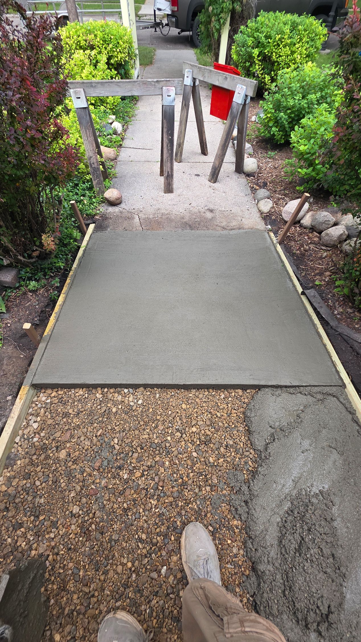 A newly poured concrete walkway section, with gravel and forms visible, next to an existing path and landscaping.